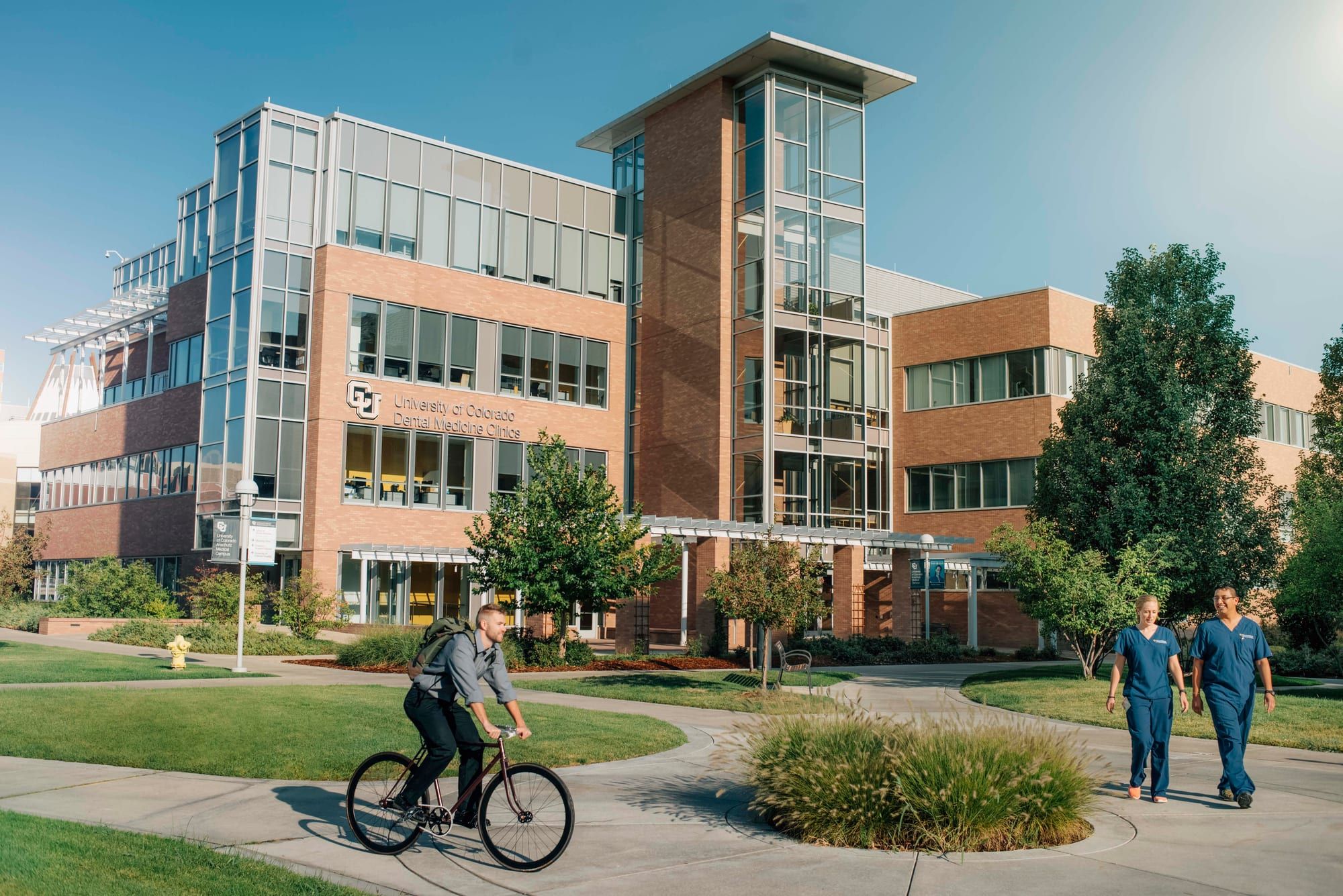 A four-story building of brick and glass with a glass stair tower, trees in front and a paved path where people walk and one person rides a bicycle